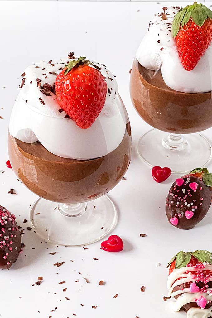 glasses of chocolate mousse on a white surface with chocolate covered strawberries and sprinkles