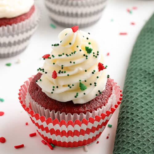 Red Velvet Cupcakes with white frosting and festive green and red sprinkles, nestled in a red and white striped wrapper beside a green textured cloth.