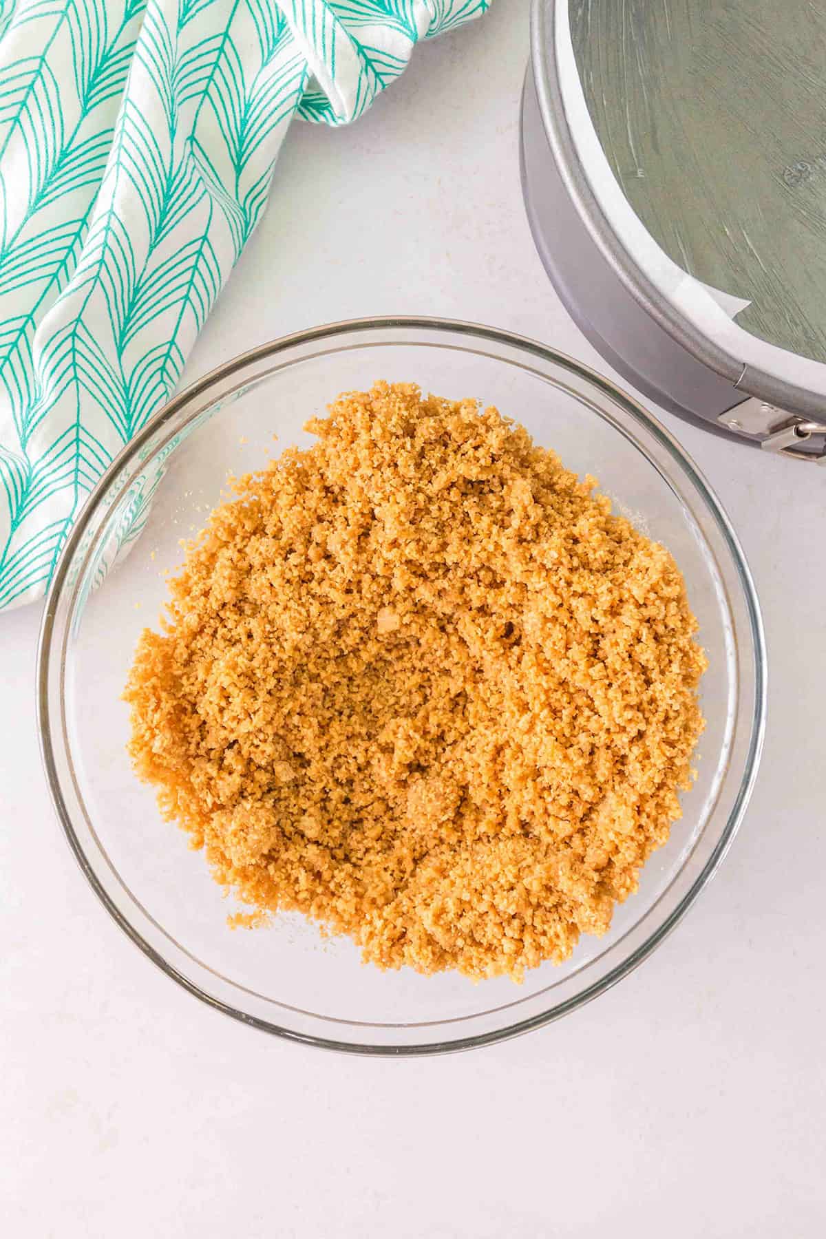 A glass bowl filled with a crumbly graham cracker crust mixture sits on a white countertop next to a springform pan and a green and white cloth, ready to become the perfect base for a luscious Strawberry Swirl Cheesecake.