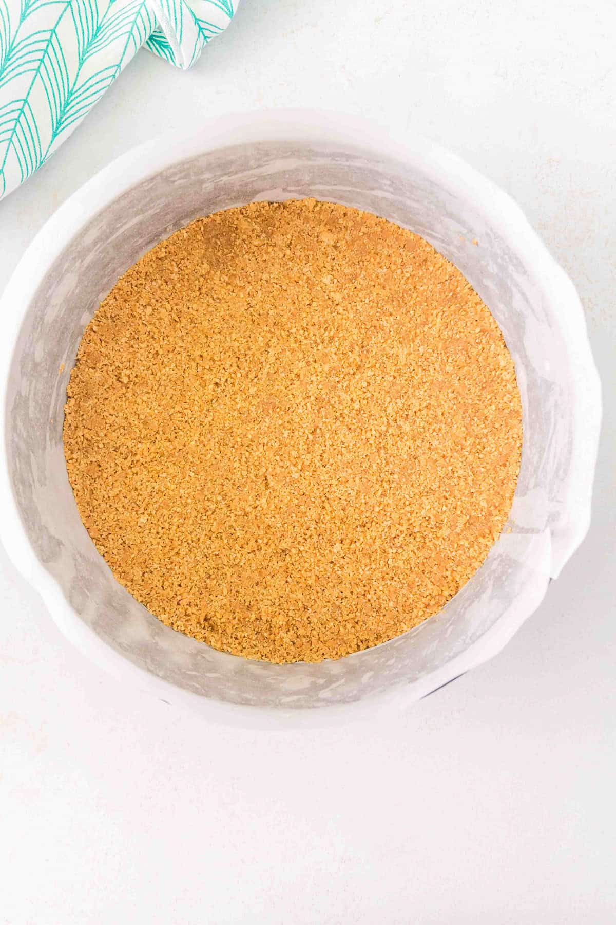 A round cake pan lined with parchment paper is filled with an even layer of graham cracker crust, forming the perfect base for a luscious Strawberry Swirl Cheesecake.