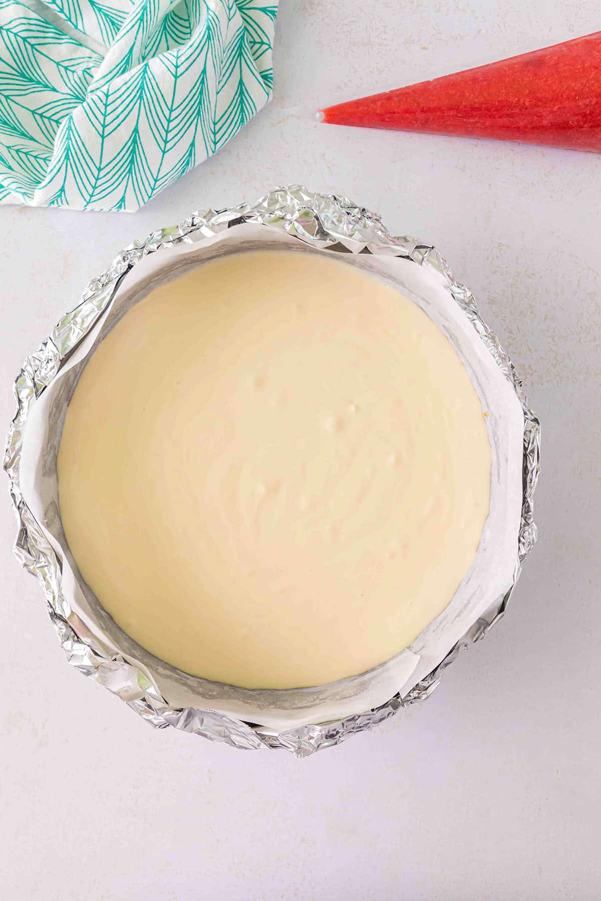 A Strawberry Swirl Cheesecake in a baking pan lined with foil sits on a light surface, with a green patterned cloth and a red piping bag nearby.