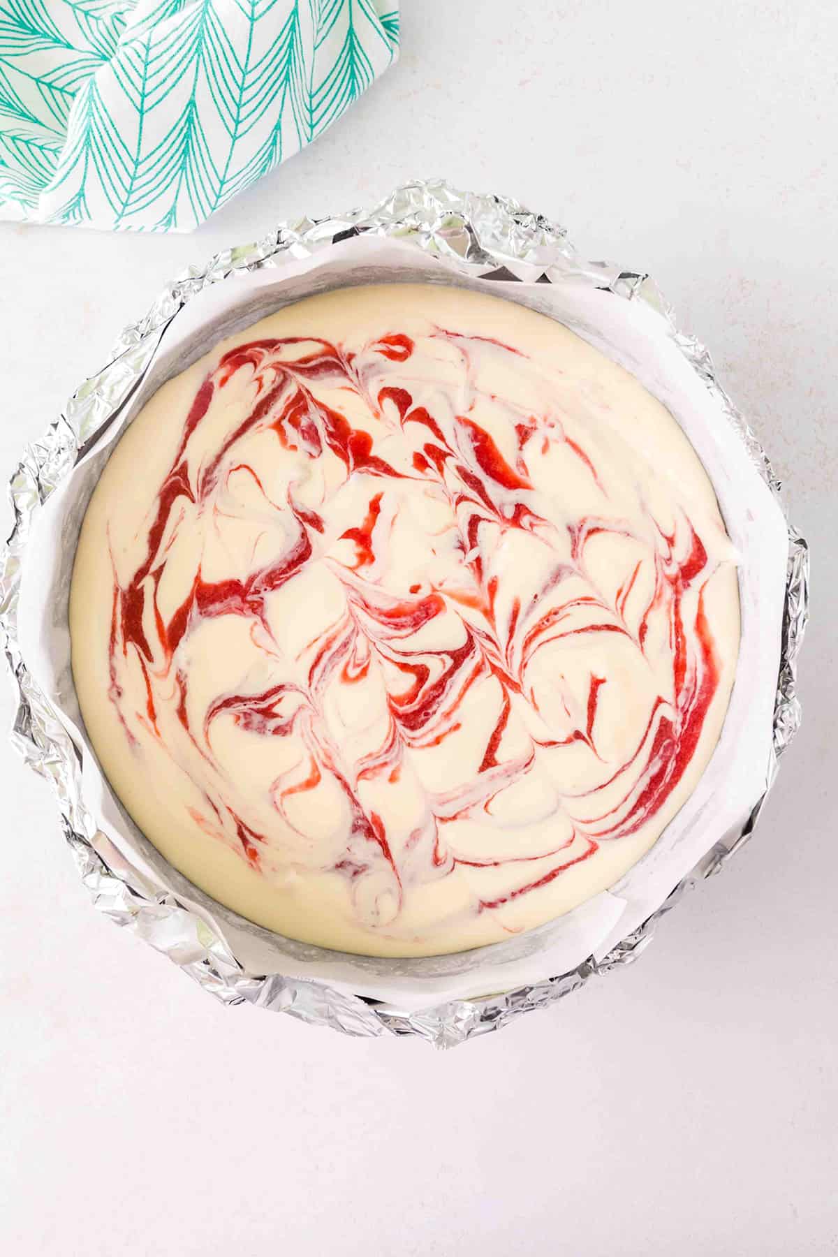 Strawberry Swirl Cheesecake batter is poured into a round, foil-lined pan and topped with vibrant red strawberry swirls in a marbled pattern, displayed on a white surface with a green patterned cloth nearby.