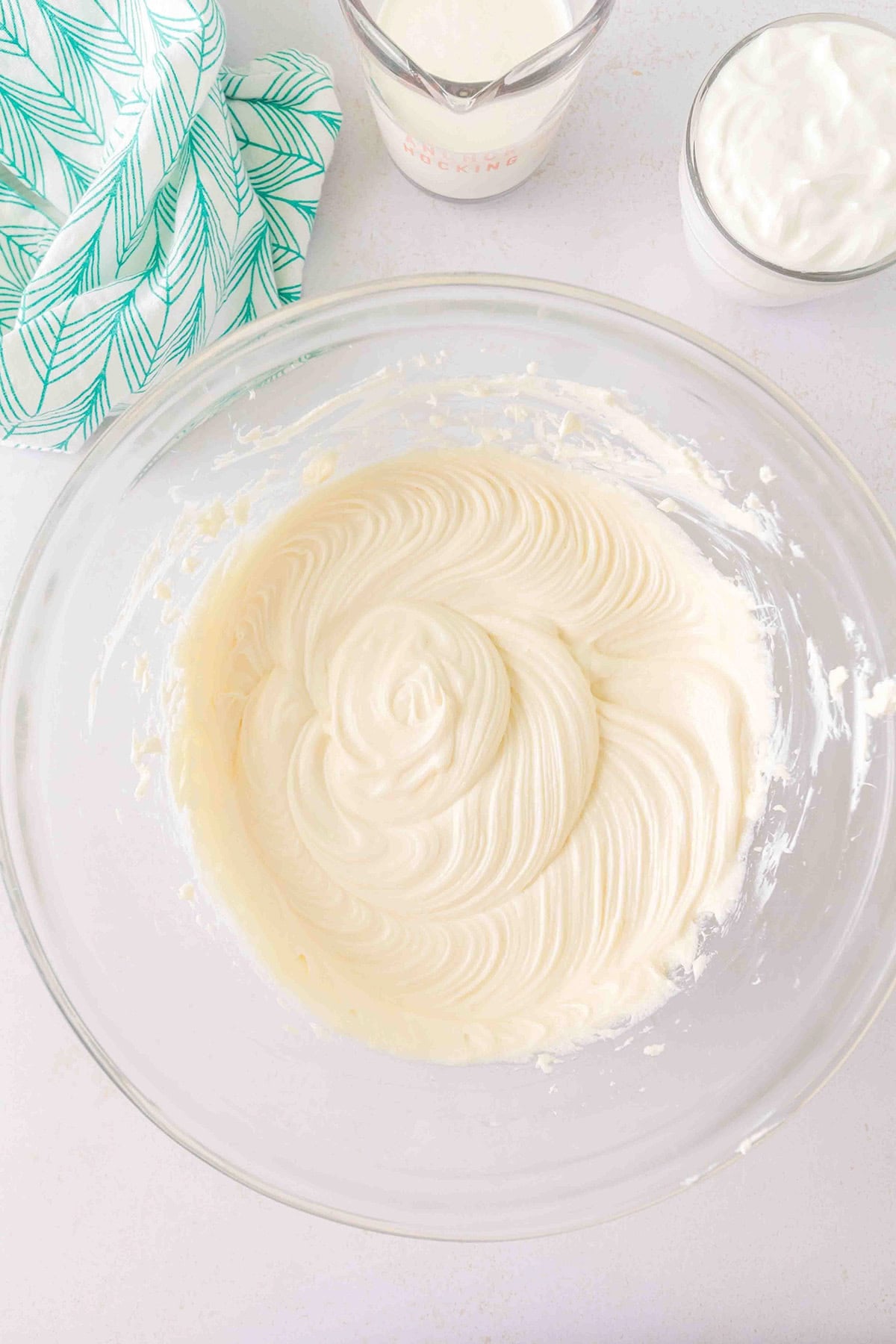 A glass bowl with thick, creamy batter sits on a white surface next to a green-striped towel, a glass of yogurt, and a measuring cup of milk—ready for creating the perfect Strawberry Swirl Cheesecake.