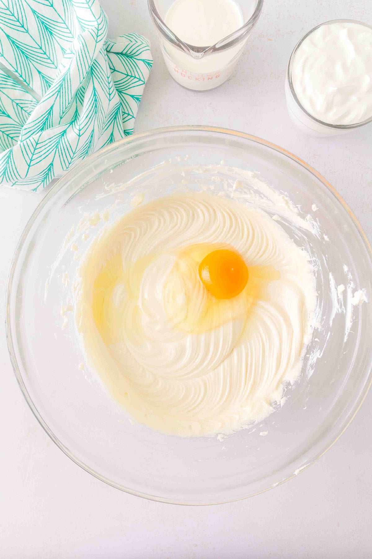 A glass bowl with creamed mixture and a raw egg, ready to become the base for a luscious Strawberry Swirl Cheesecake, sits next to a patterned towel, a measuring cup of milk, and a bowl of sour cream on a white surface.
