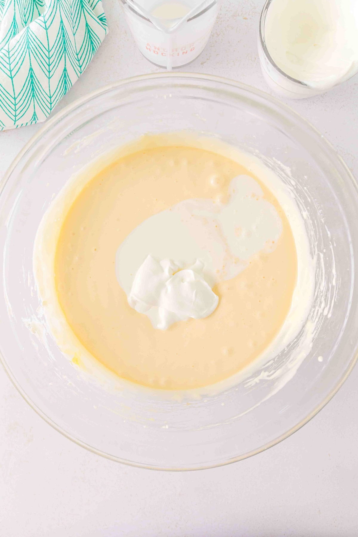 A glass bowl containing creamy batter with a dollop of sour cream on top, ready to be transformed into a luscious Strawberry Swirl Cheesecake, is surrounded by a measuring cup, glass, and patterned cloth on a light surface.