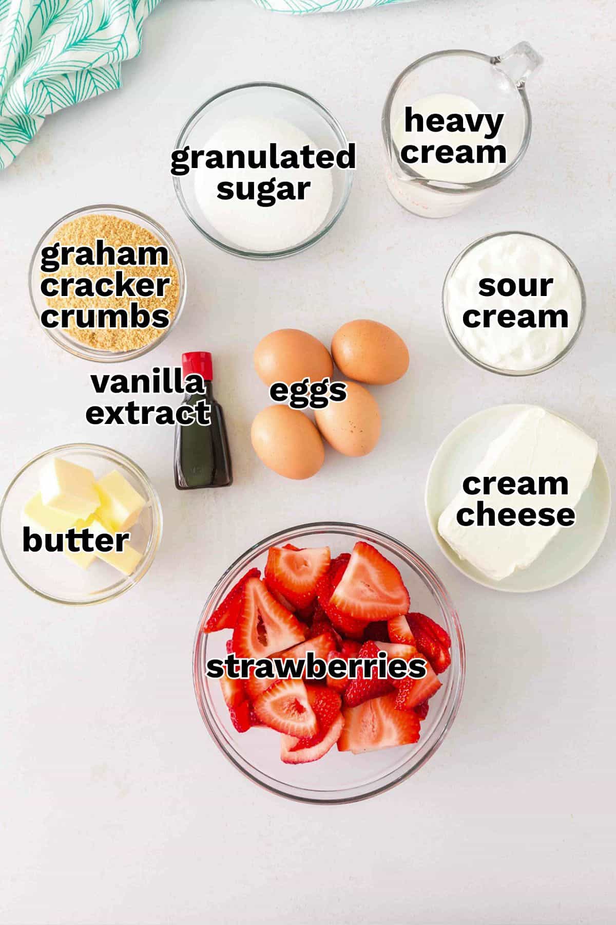 Ingredients for a Strawberry Swirl Cheesecake, including strawberries, eggs, butter, vanilla extract, graham cracker crumbs, granulated sugar, heavy cream, sour cream, and cream cheese, arranged on a white surface.
