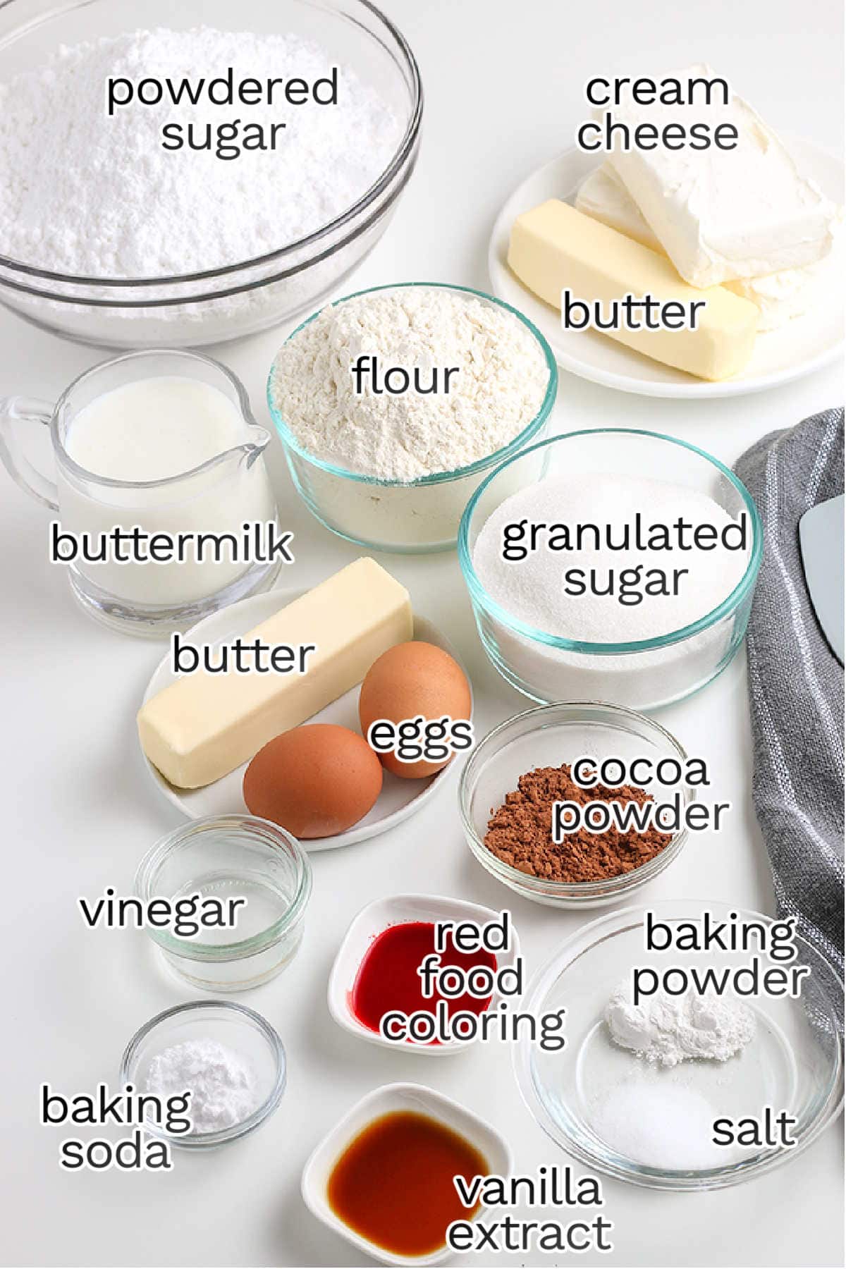 An assortment of baking ingredients in bowls and containers, including those needed for Red Velvet Cupcakes—flour, sugar, eggs, butter, cream cheese, cocoa powder, buttermilk, and flavorings—all labeled for identification.