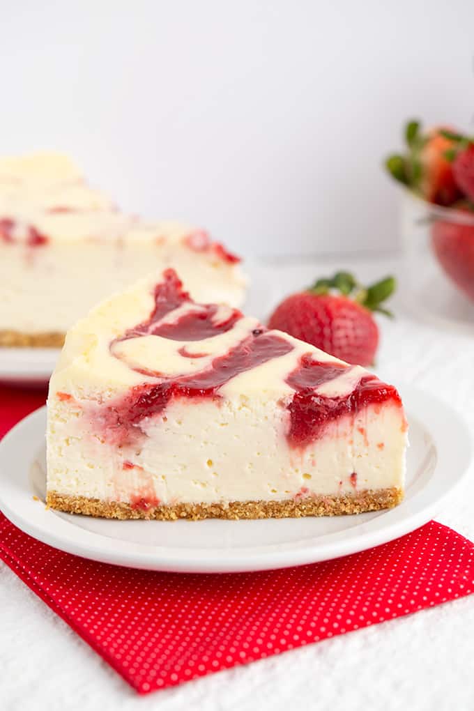 Strawberry cheesecake on a white plate with a red fabric under it and strawberries behind it