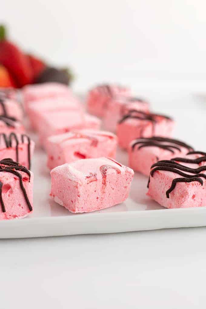 strawberry flavored marshmallows on a white tray with strawberries behind the marshmallows