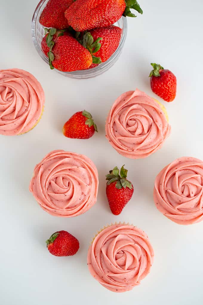 overhead photo of strawberry cupcakes on a white surface with strawberries scattered