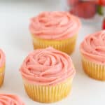 Cupcakes on a white surface with strawberries behind the cupcakes