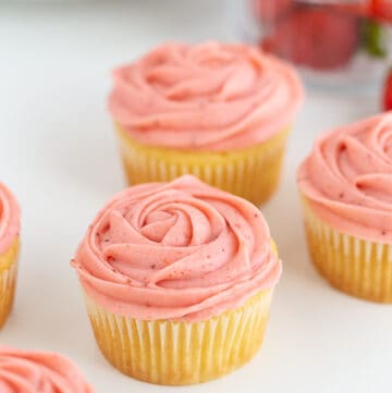 Cupcakes on a white surface with strawberries behind the cupcakes