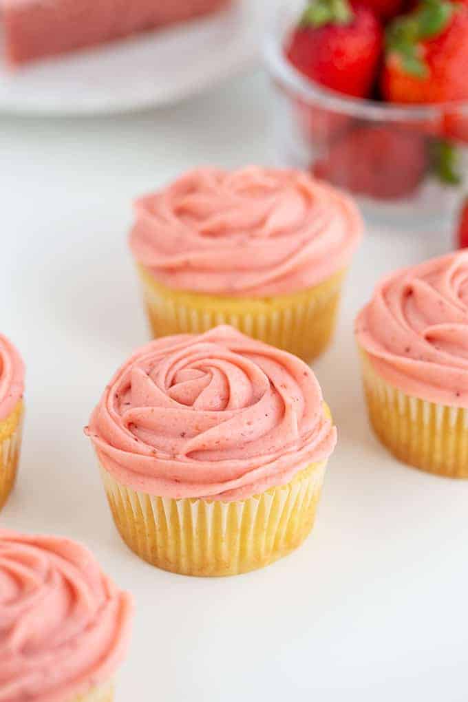 Cupcakes on a white surface with strawberries behind the cupcakes