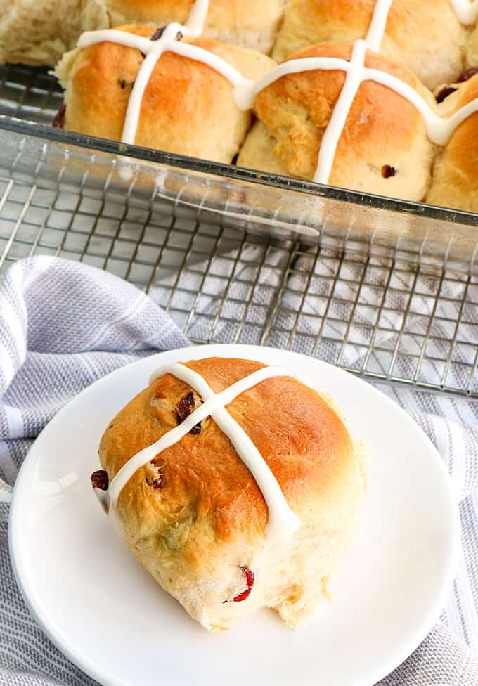 a sweet bun on a small white plate with a linen under it and a pan of buns in a baking dish beside the buns