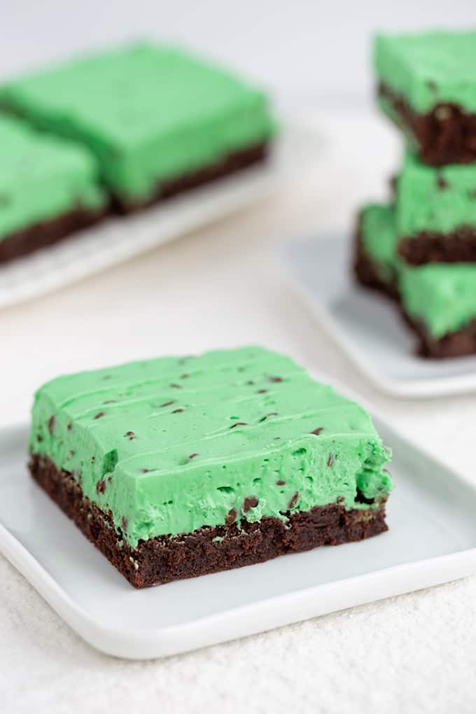 a brownie on a white square dessert plate with a stack of brownies behind in on a white surface