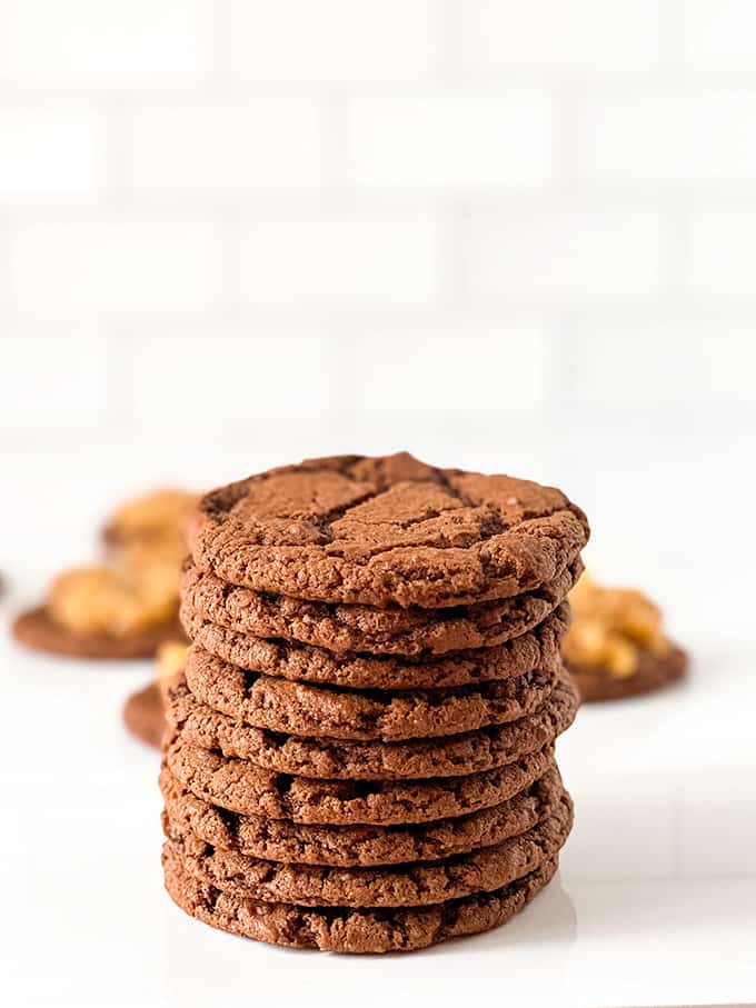 stack of chocolate cookies on a white surface with other cookies with topping behind the stack