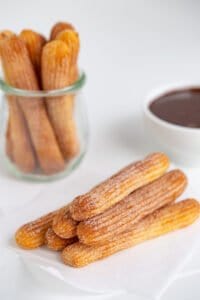side view of the stacked air fryer churros on parchment paper with a jar of churros and a bowl of chocolate sauce behind it