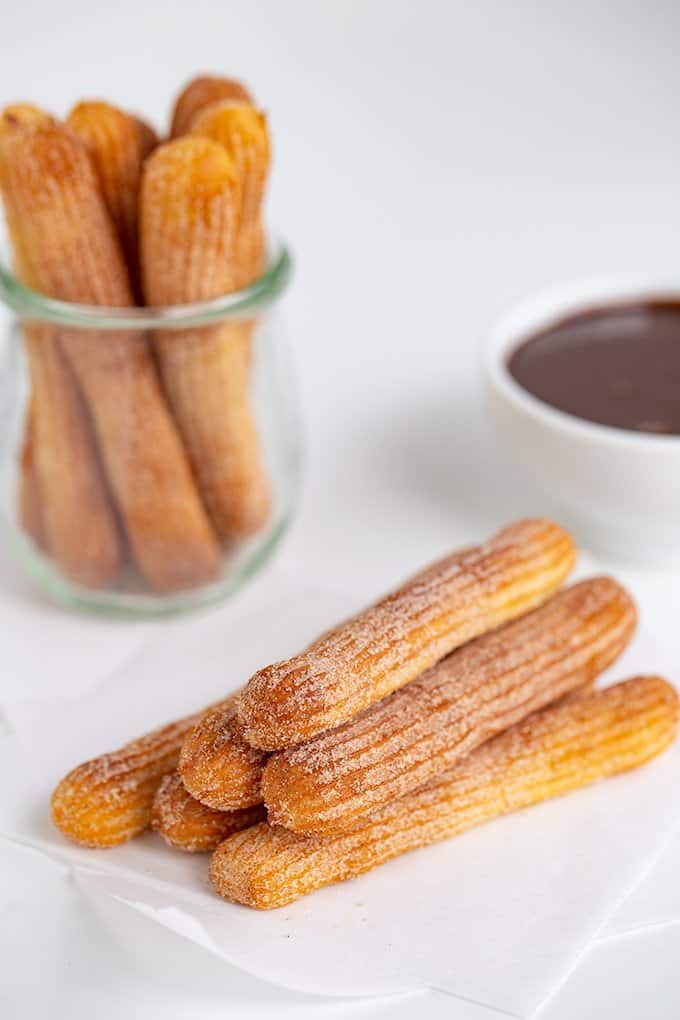 side view of the stacked air fryer churros on parchment paper with a jar of churros and a bowl of chocolate sauce behind it