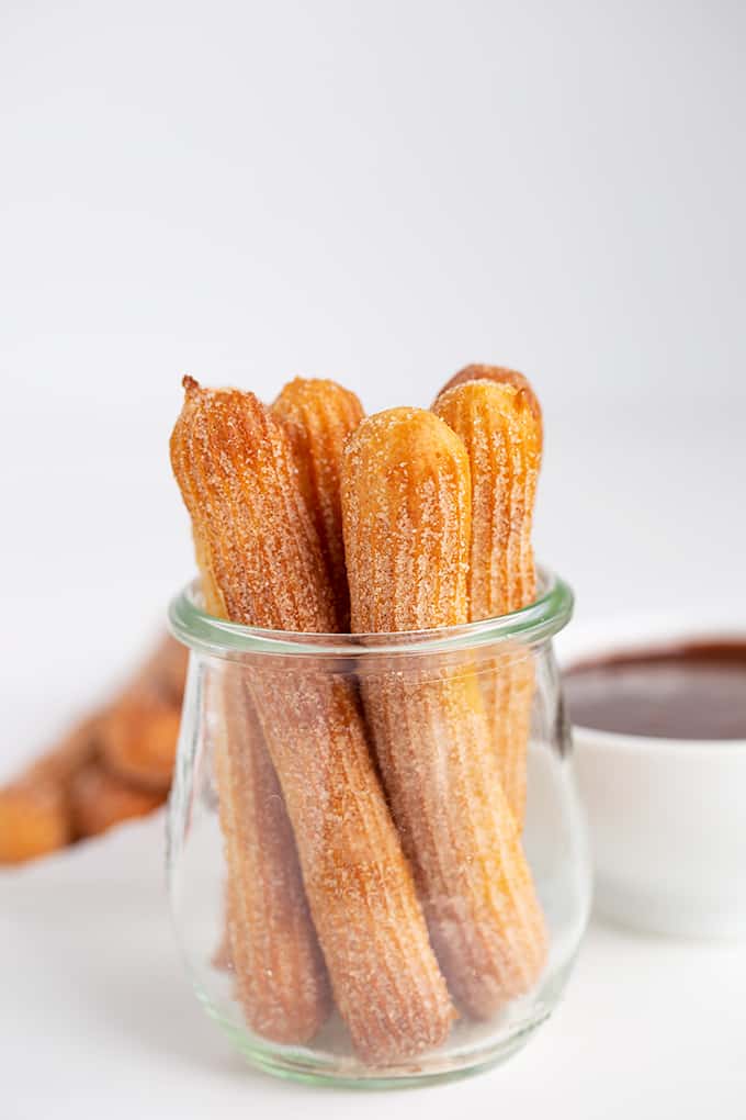 small glass jar with churros in it with dipping sauce in a white bowl behind it