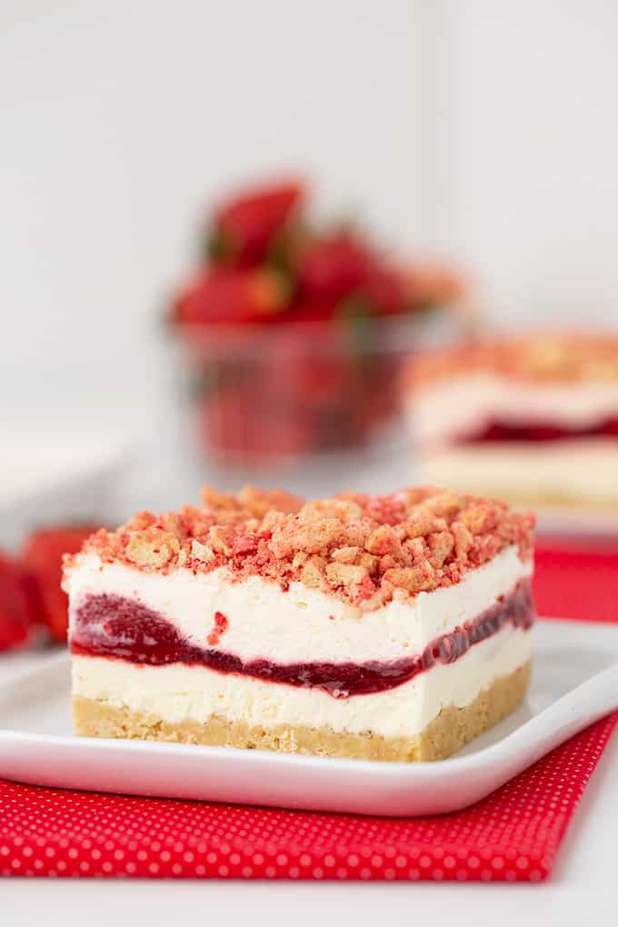 slice of dessert on a square plate sitting on a red fabric with strawberries in the background