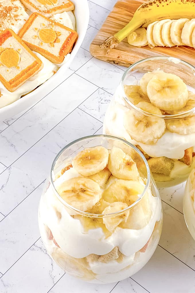 glasses of banana pudding on a white background with a casserole pan and cutting board behind it