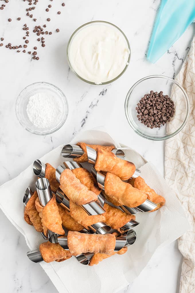 overhead photo of cannoli shells sitting on a paper towel with the ingredients for the filling around the shells