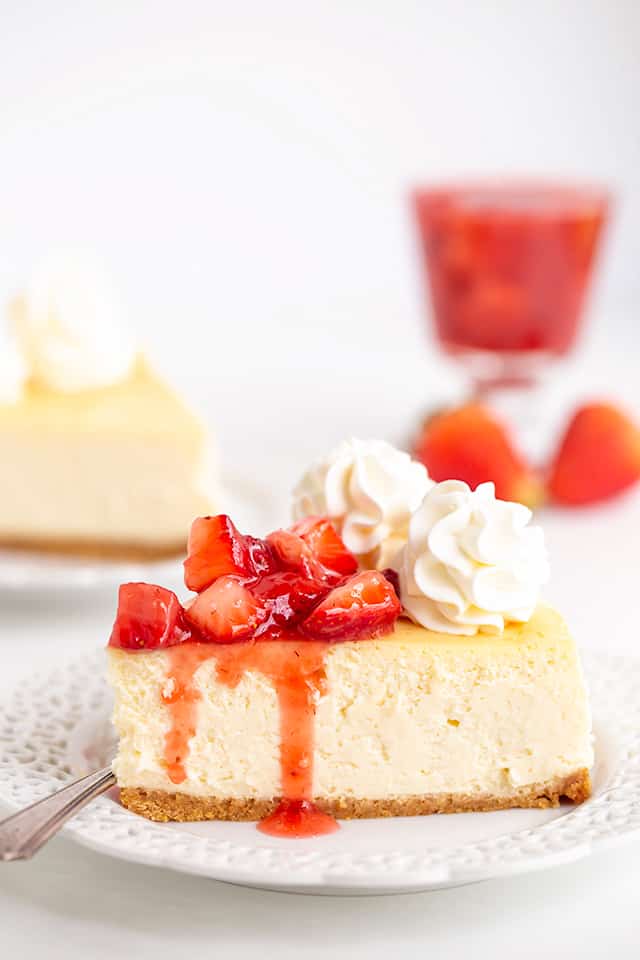 image of a slice of cheesecake on a white plate with a fork and strawberry sauce dripping down the cheesecake
