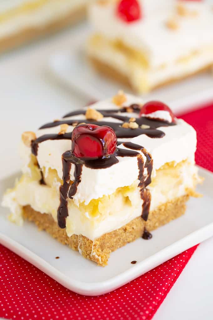 slice of no bake banana split cake on a small white plate with a red fabric under it and chocolate syrup dripping down the dessert