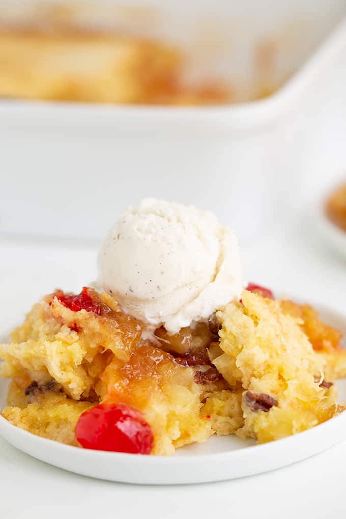 dump cake with ice cream on top on a small white plate with a white baking dish behind it