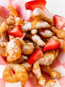 close up of funnel cake fries on a checked parchment paper with strawberries and powdered sugar