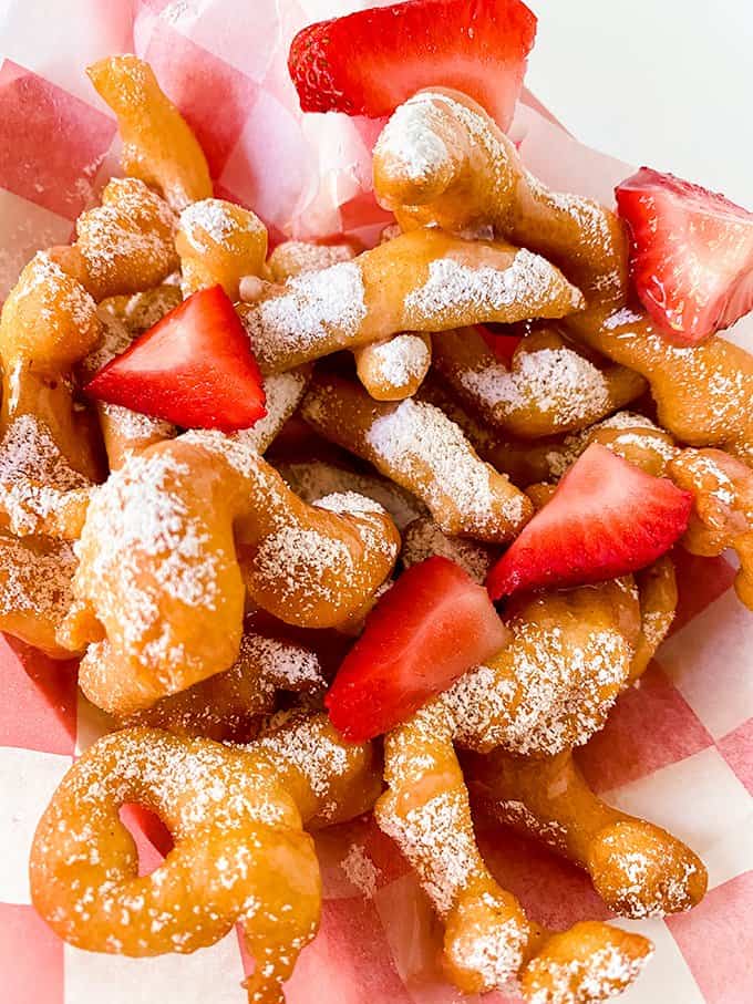 close up of funnel cake fries on a checked parchment paper with strawberries and powdered sugar