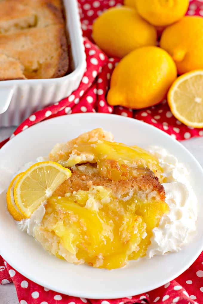 lemon cobbler on a white plate with fresh slice of lemon and whipped cream with a red and white polkadotted fabric under the plate