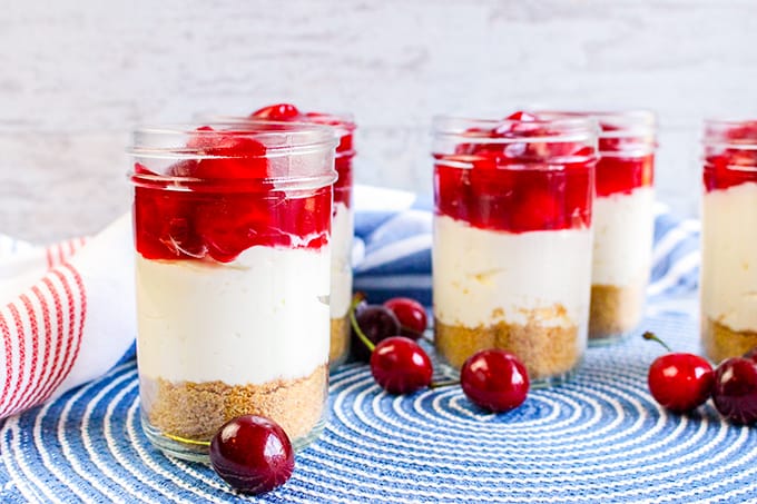 horizontal image of cheesecakes on a blue placement with cherries all around the jars of cheesecake