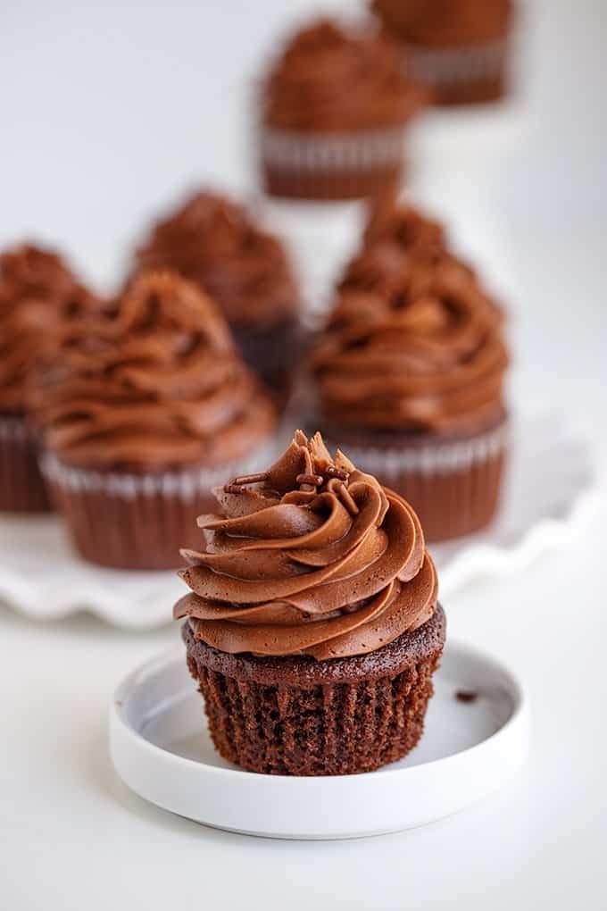 a small white plate with a cupcake that's been unwrapped and a platter of cupcakes behind it on a white surface