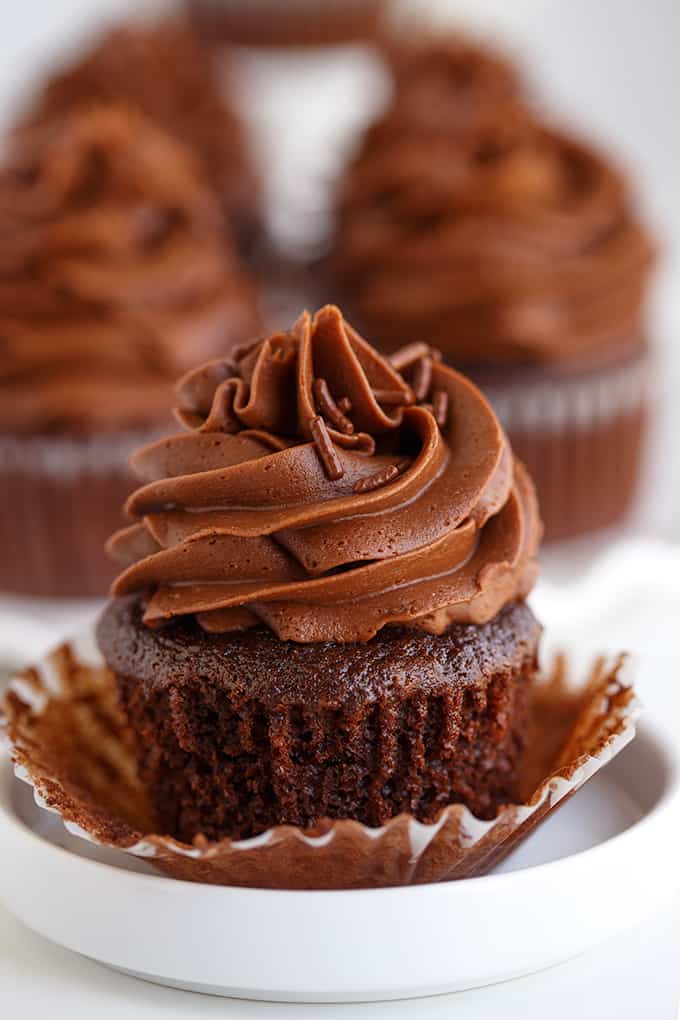 unwrapped chocolate cupcake on a small white plate with other chocolate cupcakes behind it