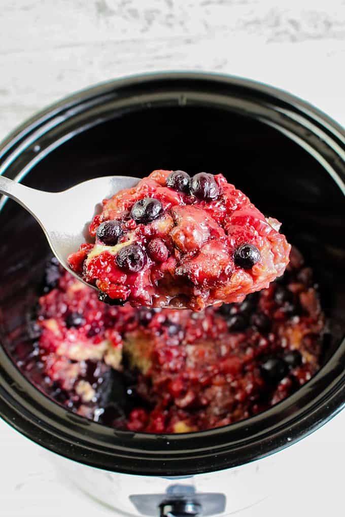 slow cooker with a black insert filled with berry cobbler and a spoon holding some above the crockpot