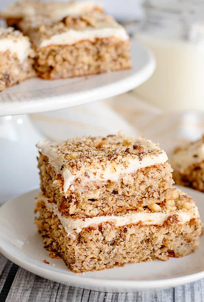 banana bars stacked on a white plate with a white cake plate behind them with more bars