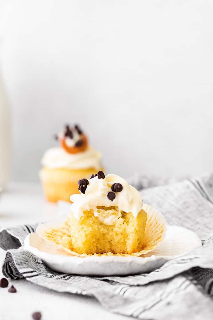 cannoli cupcakes unwrapped with a bite taken out of it on a white plate with a second cupcake behind it