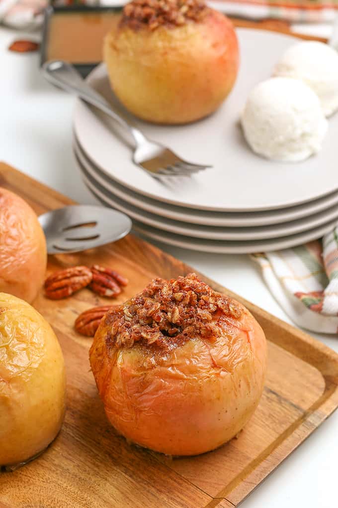 baked apples on a cutting board with a stack of white plates behind it with an apple on it