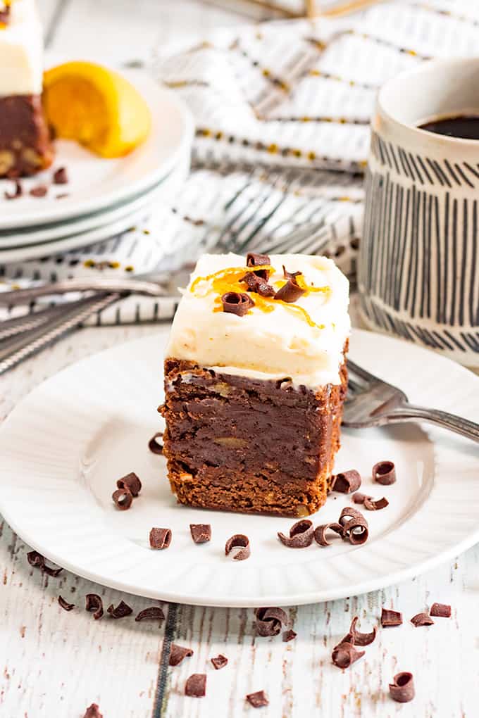 tall piece of orange brownie on a white plate with a fork and chocolate curls with a white wood background