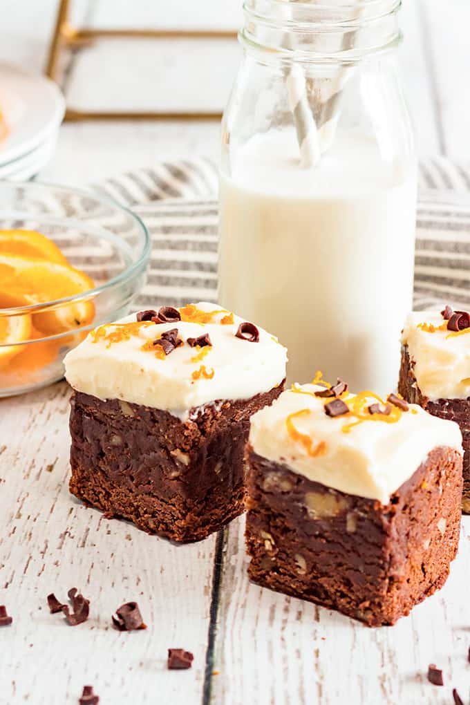 two slices of brownies on a white wooden background with a tall glass of milk with two straws behind it