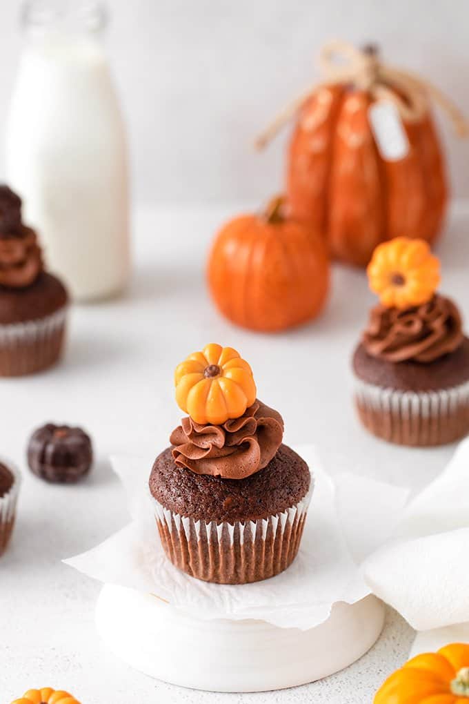 cupcakes on a white stand with parchment paper under them on a white surface and small pumpkins behind them 