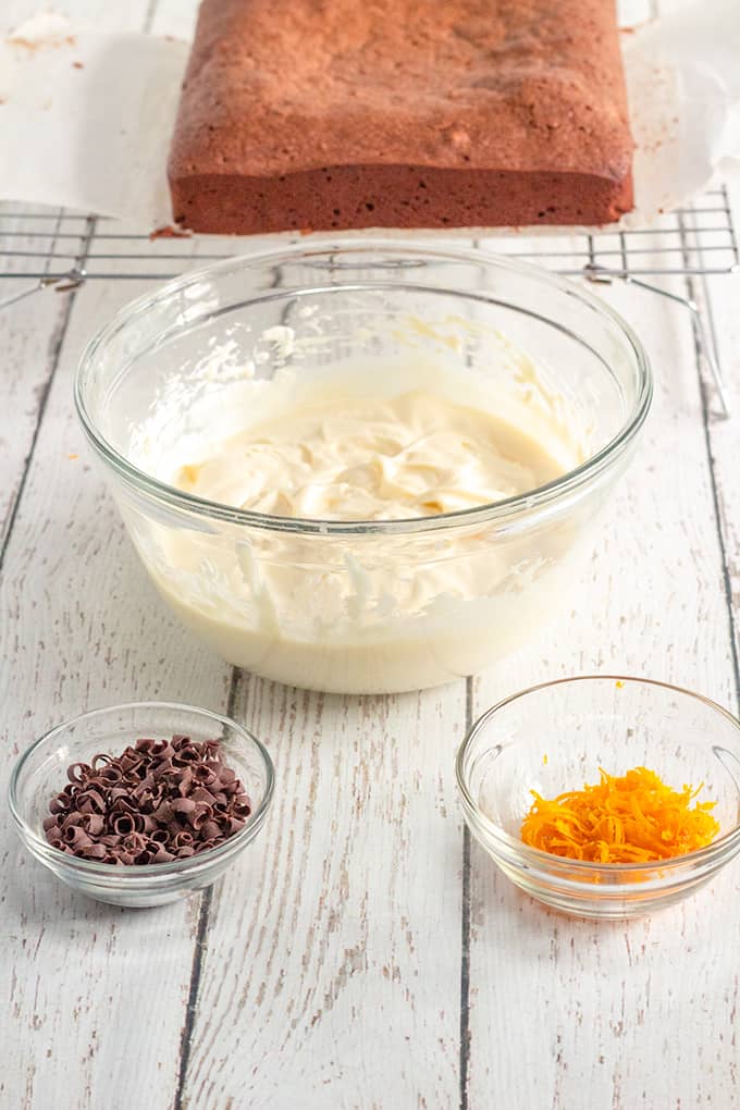 ingredients for condensed milk frosting and the toppings for the brownies in glass bowls on a wood surface with the brownie behind it