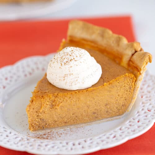 slice of pie with whipped cream and a dusting of cinnamon on top with a white lace plate and an orange linen