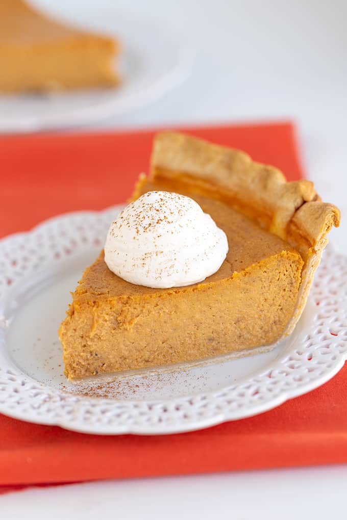 slice of pie with whipped cream and a dusting of cinnamon on top with a white lace plate and an orange linen