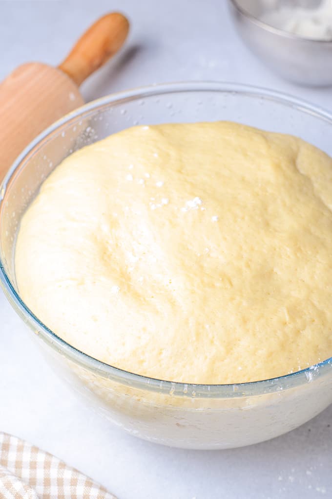 dough in a glass bowl on a white surface with rolling pin behind it