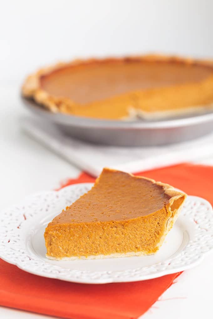 slice of pumpkin pie on a white plate with an orange fabric under it and a whole pumpkin pie behind it