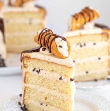 slice of vanilla cake with cannoli frosting on a white plate with the rest of the cake behind it on a white surface