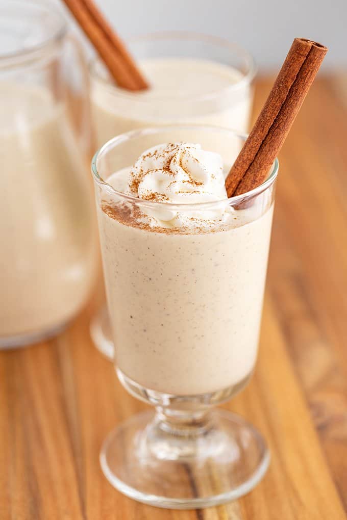 Eggnog in a glass with whipped cream, cinnamon stick, and a sprinkle of cinnamon on a wood surface