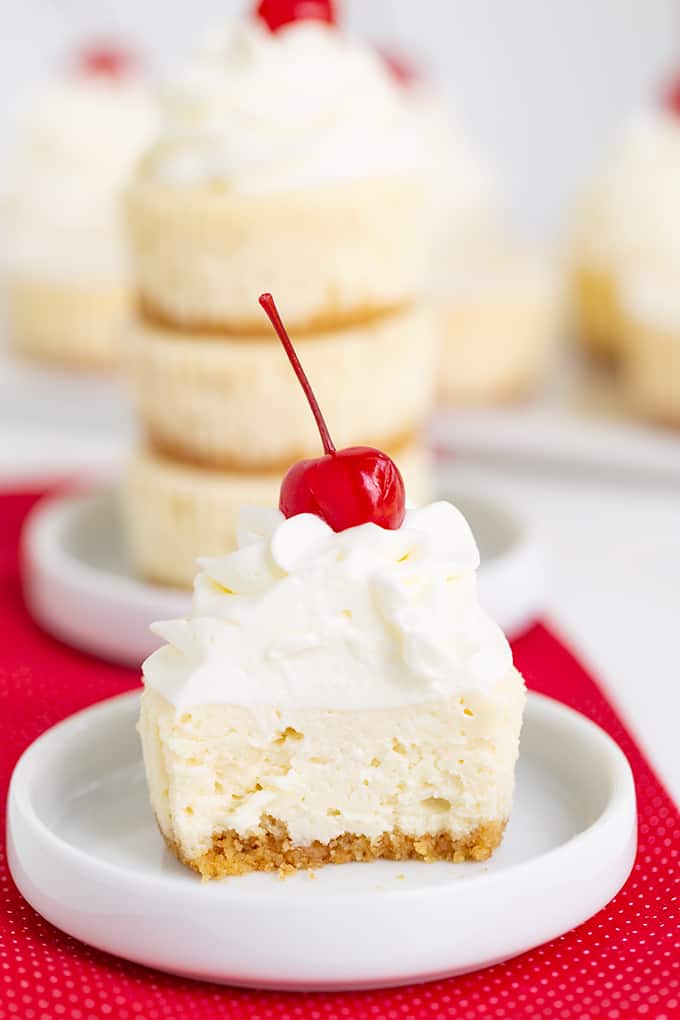 a mini cheesecake cut in half on a small white plate on a red linen and whipped cream and a stemmed maraschino cherry on top